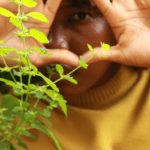 Photo of Anicka Austin wearing a yellow sweater with plants in the foreground.