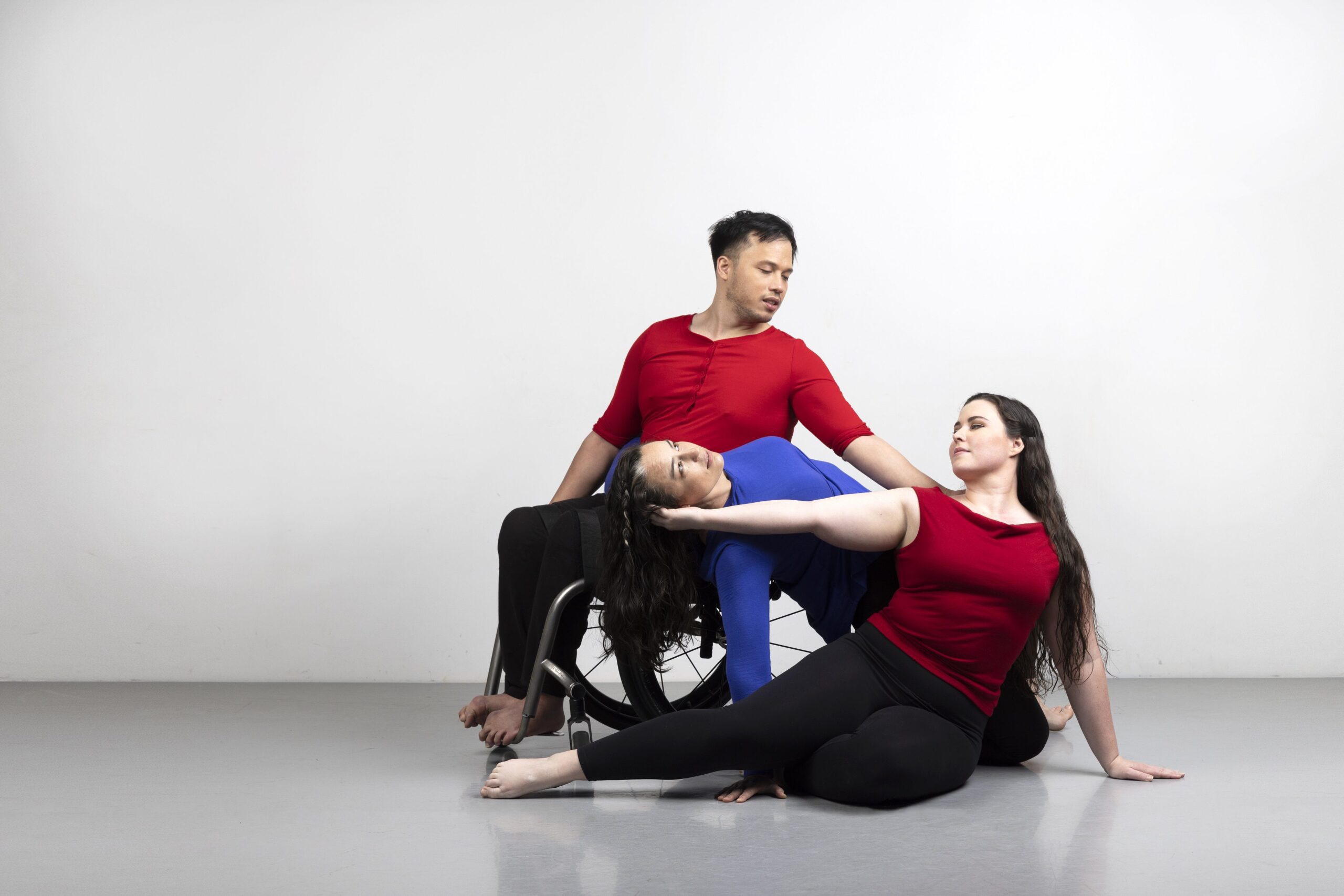 ID: Courtney, a white woman, Ashlee, a white woman, and Peter a mixed-race Asian and Caucasian man in a wheelchair, pose together. Courtney sits on the floor and cradles Ashlee’s head in her hand. Ashlee leans against Peter’s wheelchair as he reaches toward her.